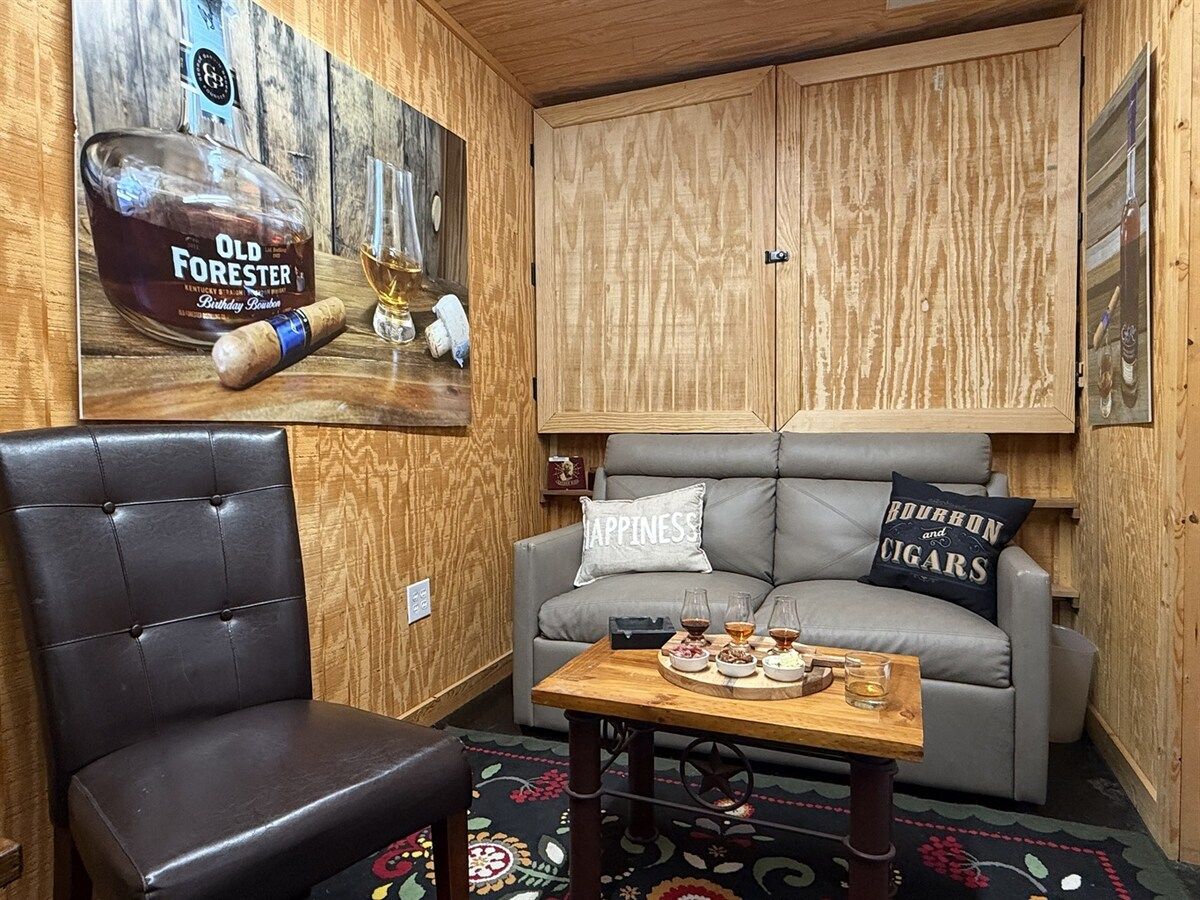 Ein gemütliches Zimmer mit einem Ledersessel, einem grauen Sofa mit Kissen, einem Holztisch mit Getränken und einem an der Wand hängenden Poster von Old Forester Bourbon.