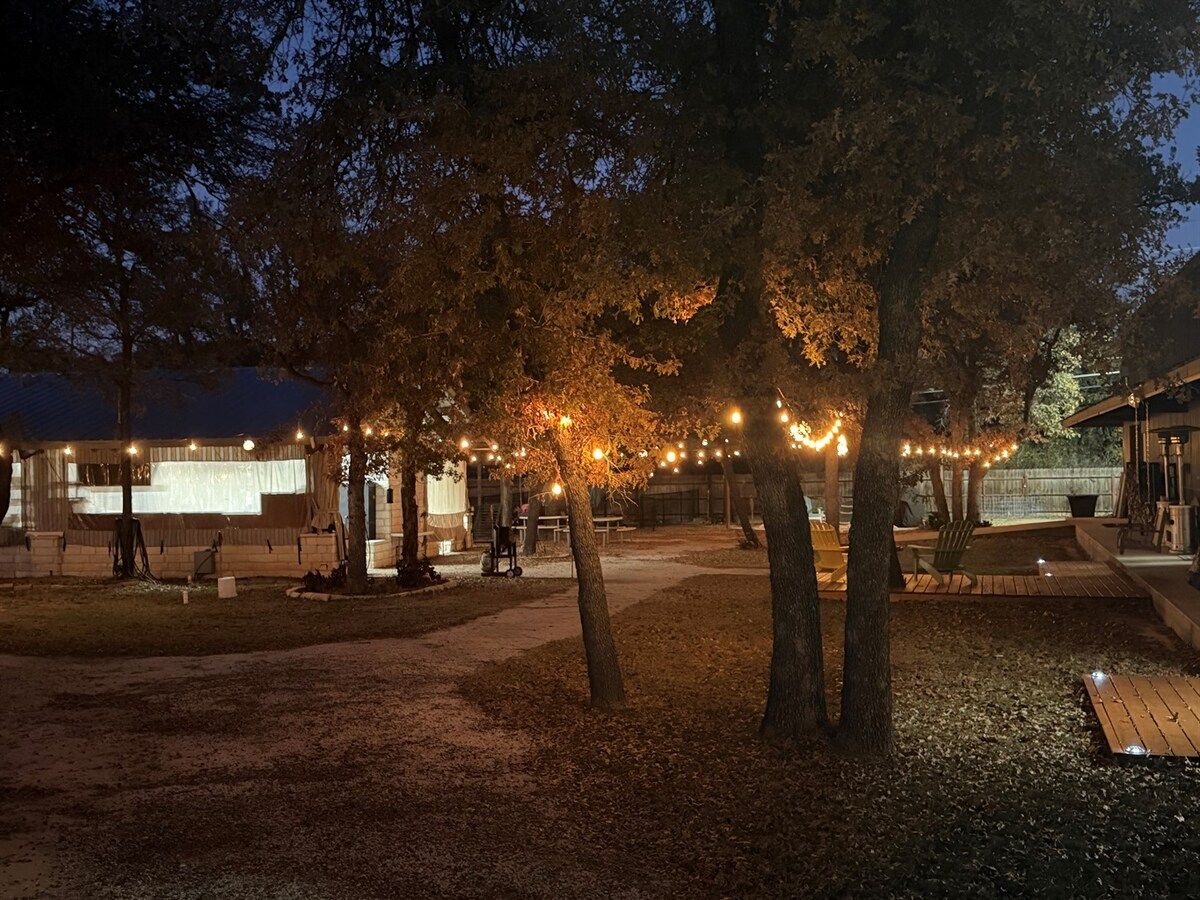 Un espacio al aire libre con árboles, una zona de estar cubierta y luces colgantes.