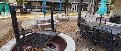 Un pozo de fuego de piedra con sillas y mesas de metal, rodeado por una terraza de madera y árboles.
