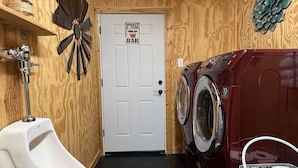 Des toilettes pour hommes avec un urinoir, une grande horloge murale, une porte portant l’inscription « Bar à whisky et cigares » et un lave-linge rouge.