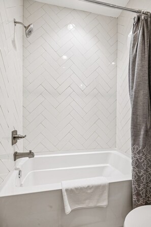 Ein modernes Badezimmer mit Wandfliesen in Fischgrätmuster, einer Dusche inklusive Handbrause sowie einer Badewanne mit Handtuch.