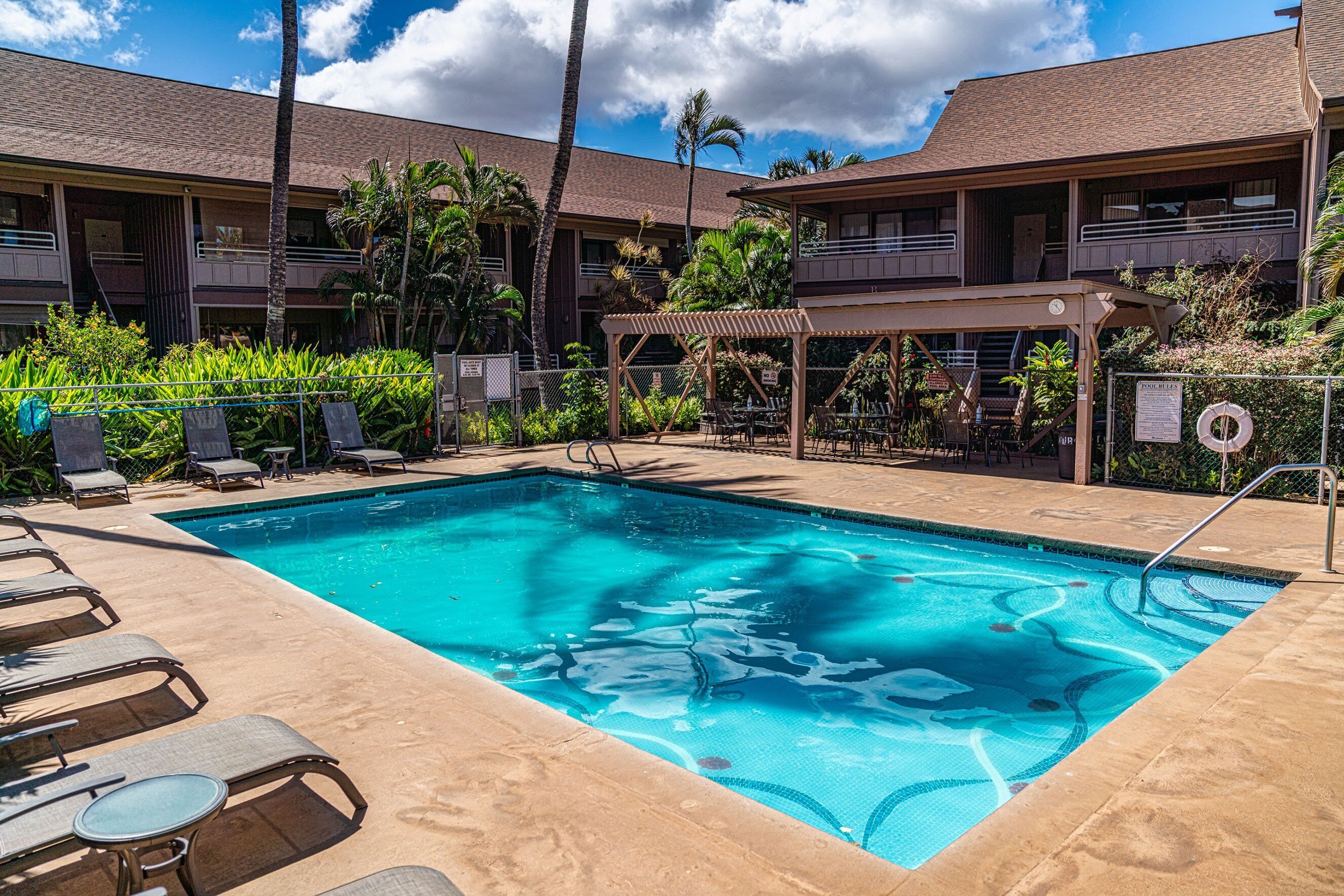 A swimming pool with lounge chairs and a covered seating area, surrounded by a building with multiple balconies and palm trees.