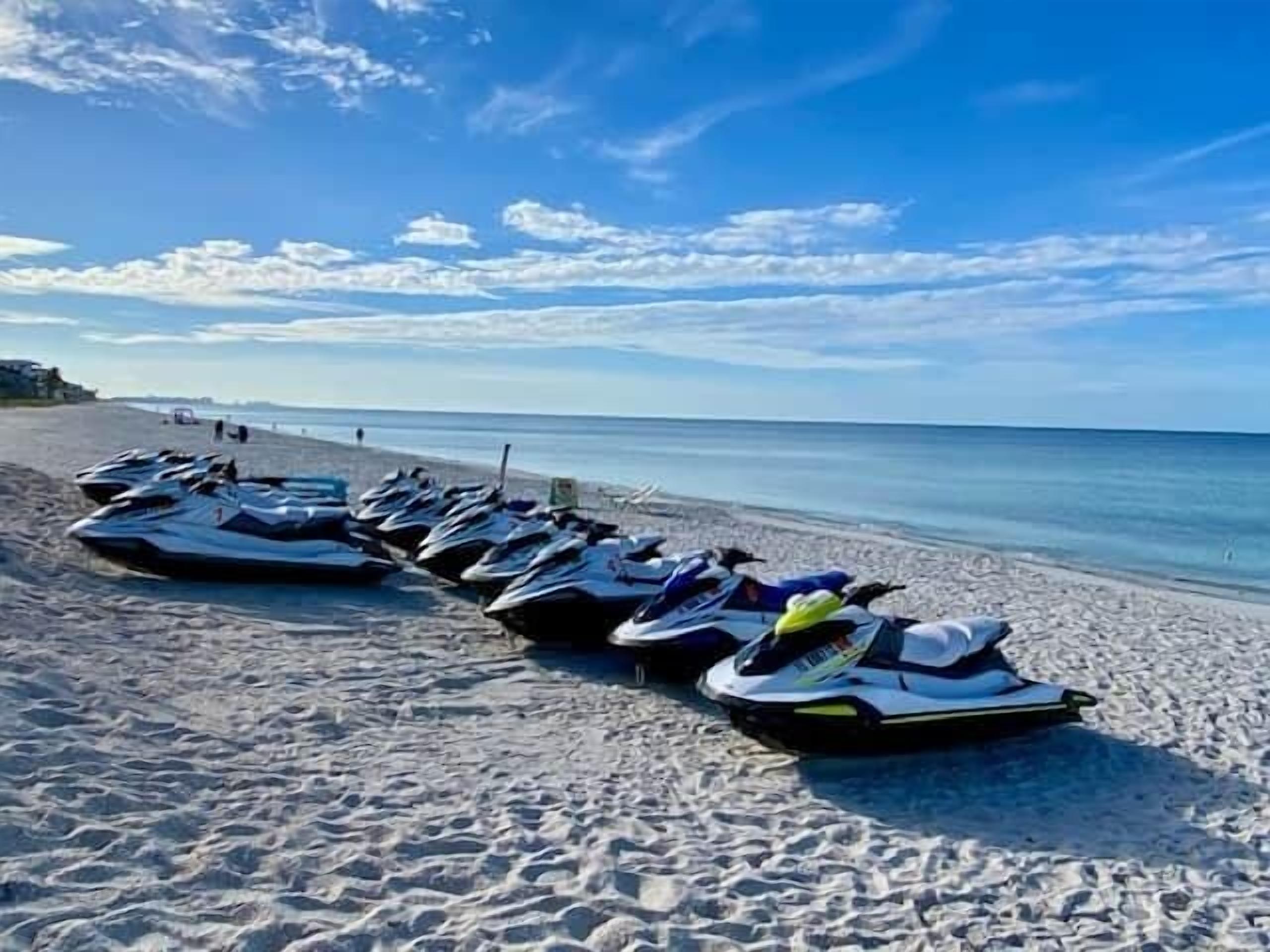 Jet skis lined up on a sandy beach with the ocean in the background.