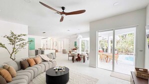 Amplia sala de estar con sofá, mesa de centro y ventilador de techo. Zona de comedor con mesa y sillas, y vista a una piscina a través de un ventanal.