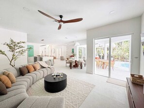 A spacious living room with a sofa, coffee table, and a ceiling fan. There is a dining area with a table and chairs, and a view of a swimming pool through a glass door. - Naples FL | Heated Pool Spa Pickleball Mini Golf (Naples)