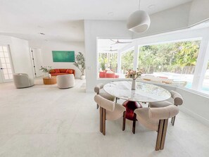 Amplia sala de estar con una mesa de vidrio redonda rodeada de sillas acolchadas, un sofá rojo y un ventanal con vistas a una piscina y vegetación.