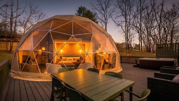 Estructura en forma de cúpula geodésica con cubierta de madera, mobiliario exterior y una iluminación interior cálida.