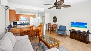 Una sala moderna con sofá, mesa de centro y televisión. Se observa una zona de cocina con armarios de madera y una mesa de comedor.