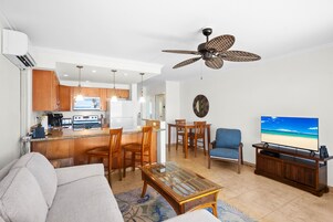 A modern living room with a sofa, coffee table, and TV. There is a kitchen area with wooden cabinets and a dining table.