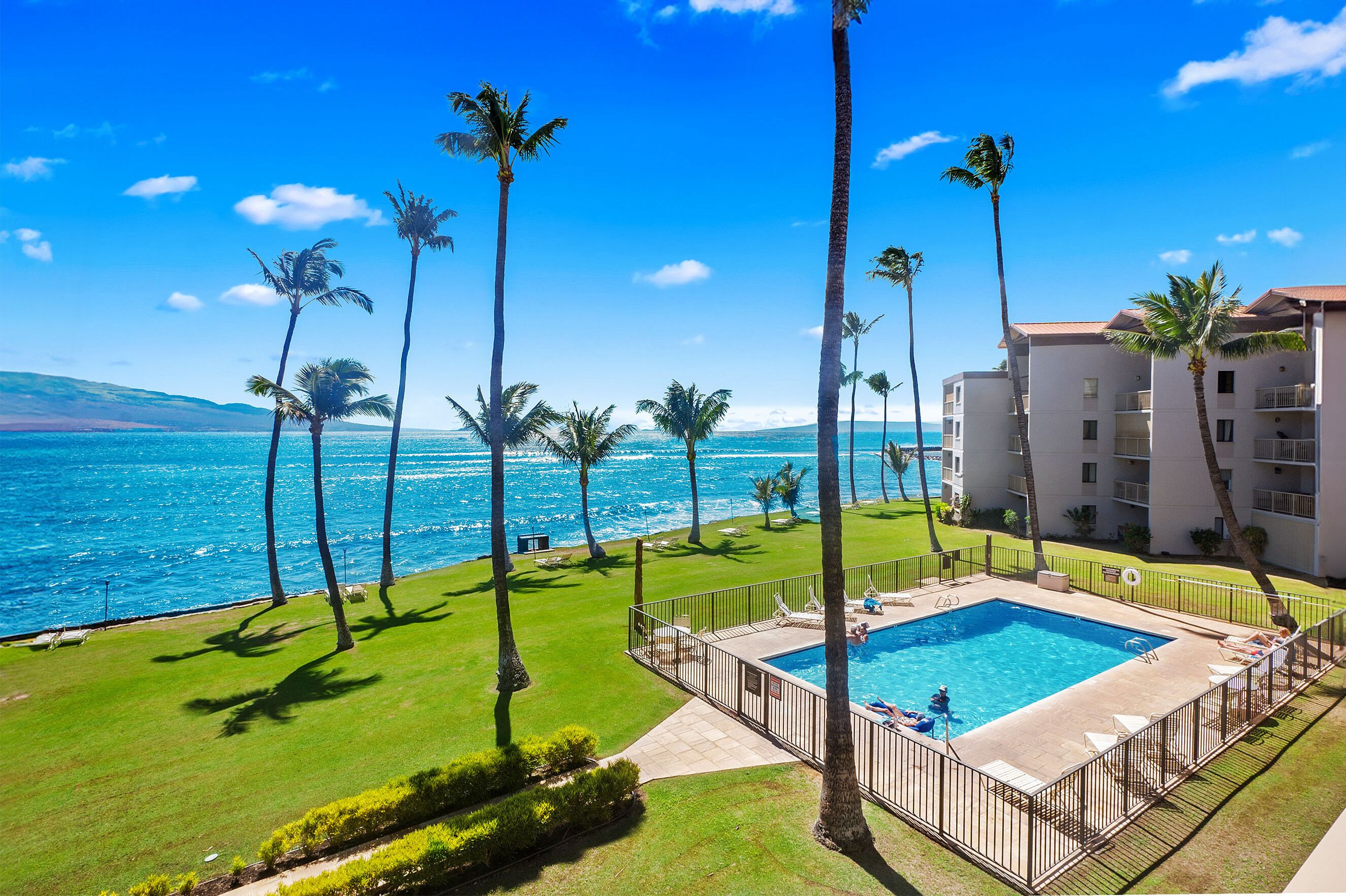 A pool area with a view of the ocean, surrounded by palm trees and a building with balconies.