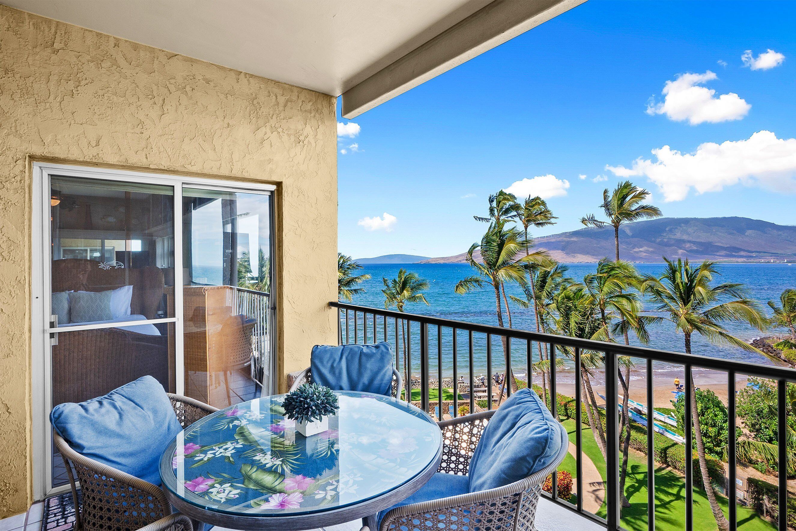 A balcony with wicker furniture and a glass table, overlooking a beach with palm trees and a mountain in the distance.