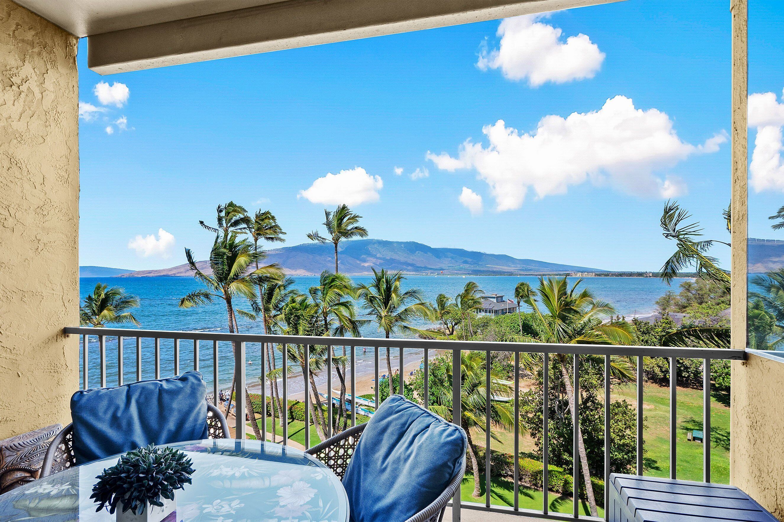 A balcony with a glass table and chairs, overlooking a beach and palm trees.