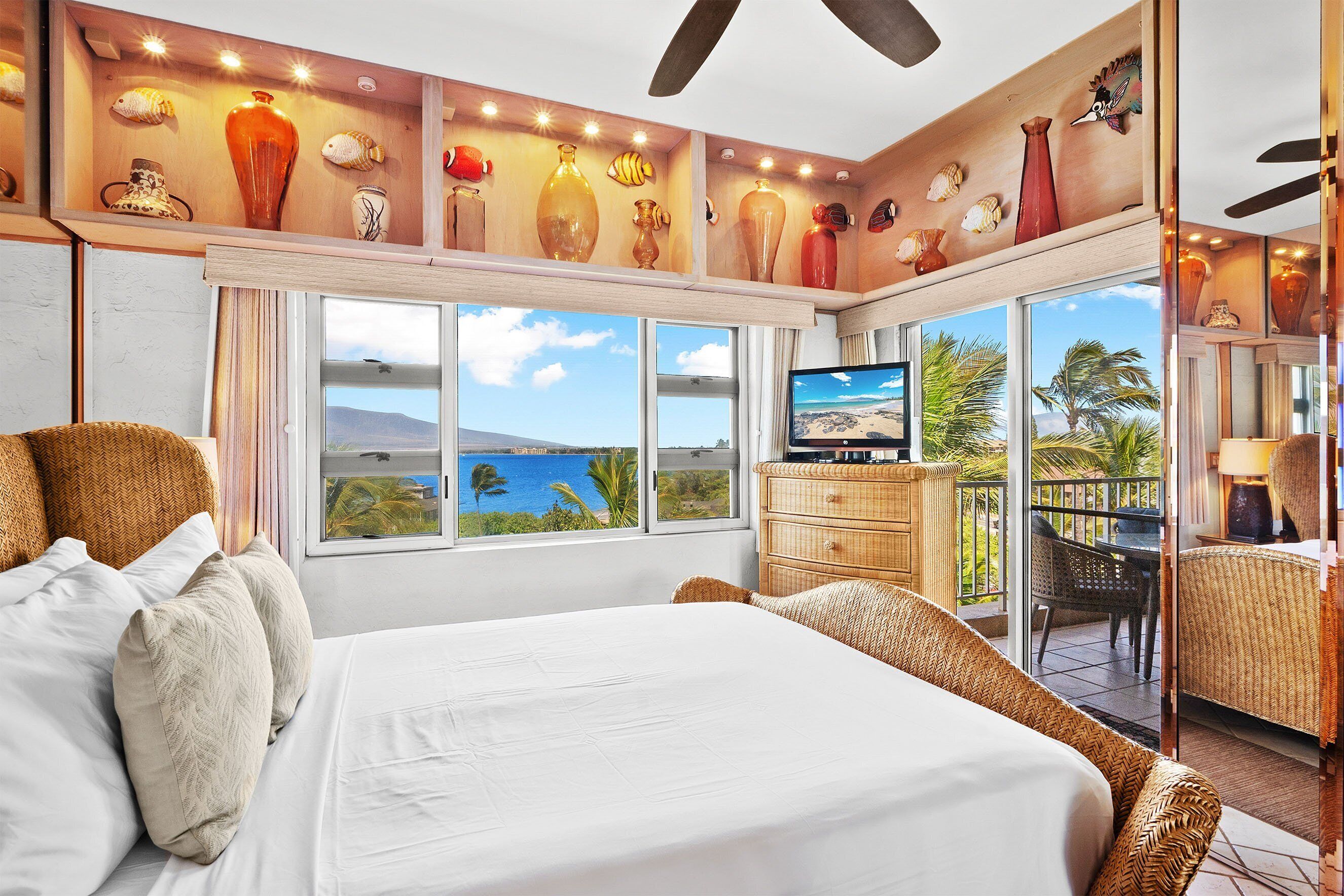 A hotel room with a large bed, a television, a balcony with a view of the ocean, and a shelf with decorative items.