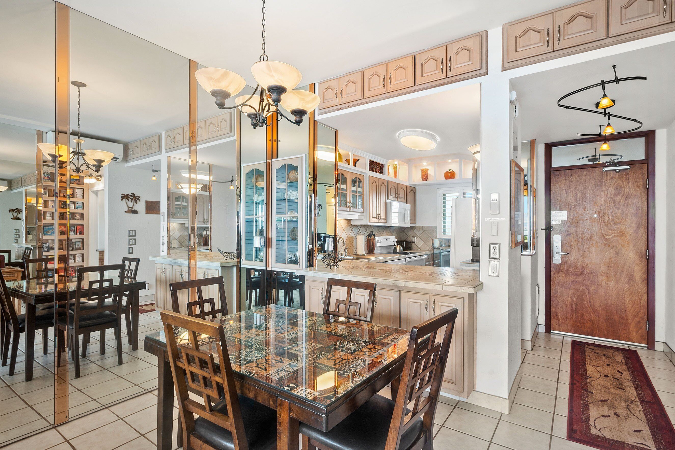 Un comedor con una mesa de vidrio, sillas de madera y un candelabro. Hay una zona de cocina con armarios de madera y una puerta de madera.