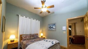 Un dormitorio con cama, dos lámparas de noche, ventilador de techo, ventana con cortinas, un cuadro enmarcado en la pared y una puerta que da a otra habitación.