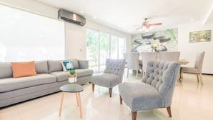 Ein modernes Wohnzimmer mit einem grauen Sofa, einem runden Couchtisch und einem gesteppten Sessel. Im Hintergrund ist ein Essbereich mit Tisch und Stühlen zu sehen.