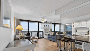 A modern living room with a dining area, a kitchen, and large windows offering a view. - Golden Sands 1513 (Ocean City)