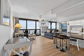 A modern living room with a dining area, a kitchen, and large windows offering a view. - Golden Sands 1513 (Ocean City)