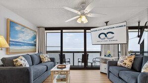 A modern living room with a large window, a ceiling fan, and a sign for Ocean City Vacation Homes. - Golden Sands 1513 (Ocean City)