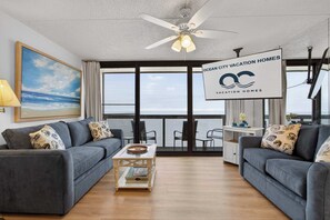 A modern living room with a large window, a ceiling fan, and a sign for Ocean City Vacation Homes. - Golden Sands 1513 (Ocean City)