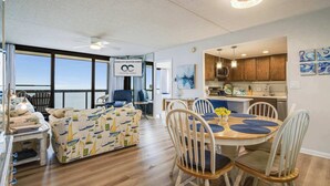 A modern living room with a large window, a sofa, a dining table with chairs, and a kitchen area. - Golden Sands 1412, Oceanfront, Pools, Games (Ocean City)