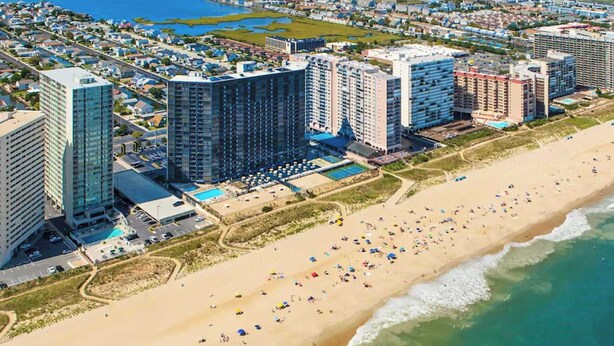 A coastal hotel area with multiple high-rise buildings, a sandy beach, and a pool area. - Golden Sands 1412, Oceanfront, Pools, Games (Ocean City)
