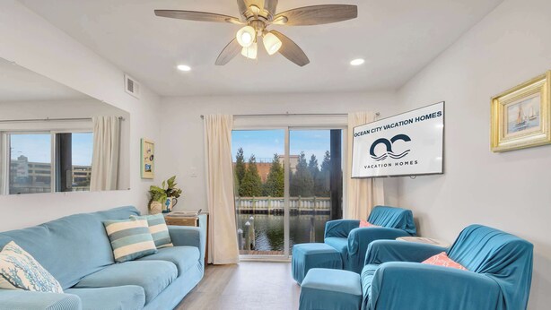 Un salon moderne avec un canapé bleu, un pouf assorti et un ventilateur de plafond. Une grande fenêtre offre une vue sur une étendue d’eau et des arbres.