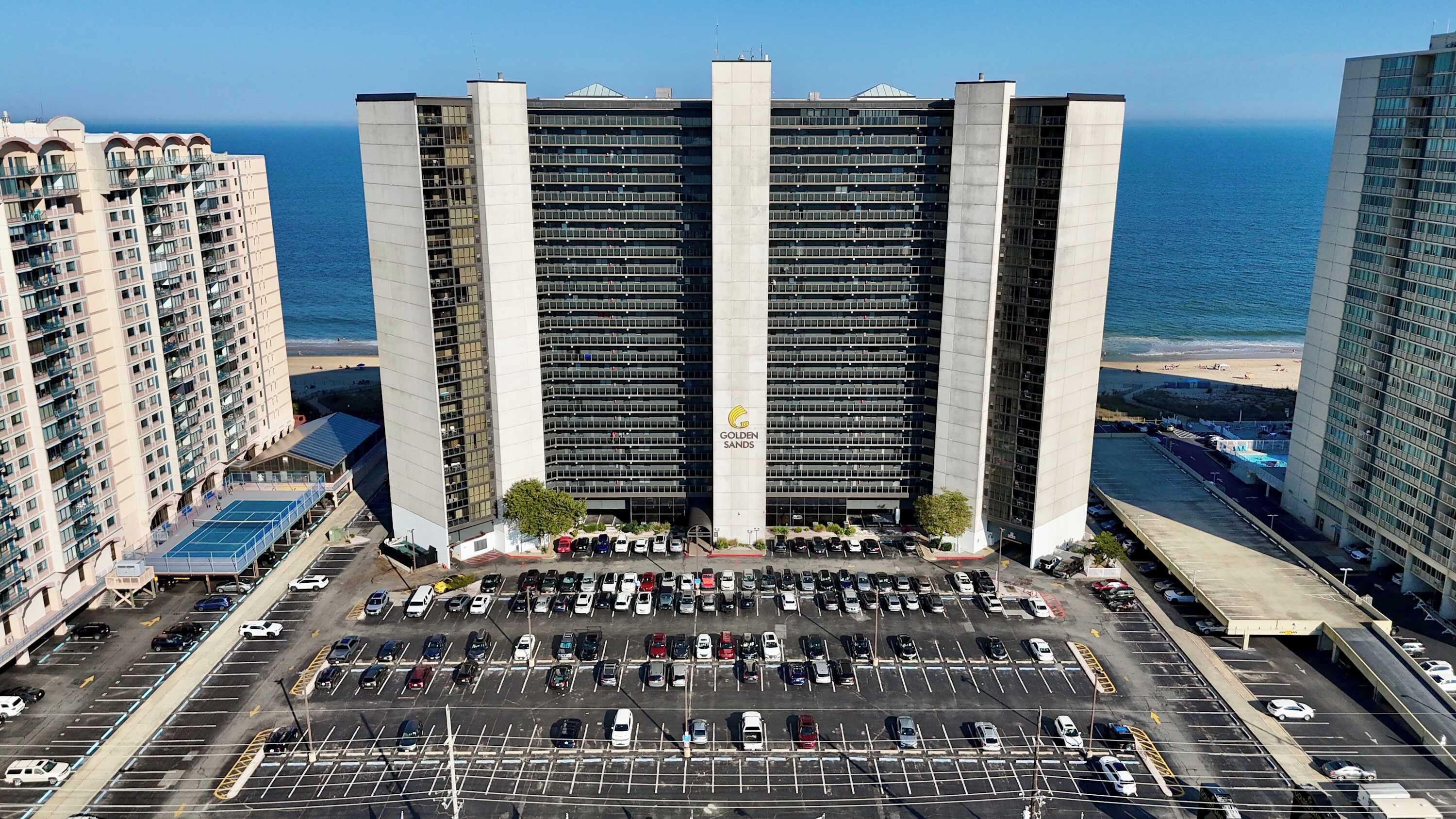 A high-rise hotel with a central tower and multiple balconies, a parking lot in front, and a beach view.