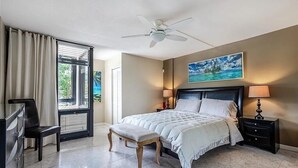 Una camera da letto con un letto, una panca, un comodino, una lampada, una ventola a soffitto e una finestra con tende.