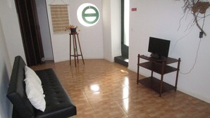 Una stanza con un divano nero, un mobiletto in legno per la TV, un piccolo sgabello con una pianta e una finestra circolare con cornice verde.
