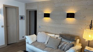 Una sala de estar con un sofá de color claro, un panel de pared de madera, dos luces empotradas, una puerta y una mesita.