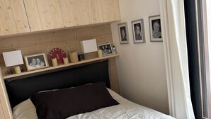 Un dormitorio con una cama, cabecera de madera, estante con fotos enmarcadas y adornos, y una ventana con cortinas.
