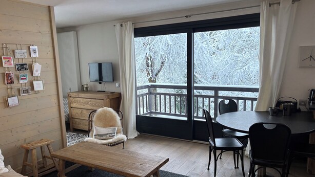Una sala de estar acogedora con una mesa de centro de madera, un sofá y un comedor con sillas. Un televisor está montado en la pared, y hay cuadros en la pared.