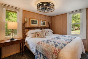 Un dormitorio con pared de paneles de madera, una cama con colcha, dos cuadros enmarcados, una lámpara y una ventana con vistas a la vegetación.