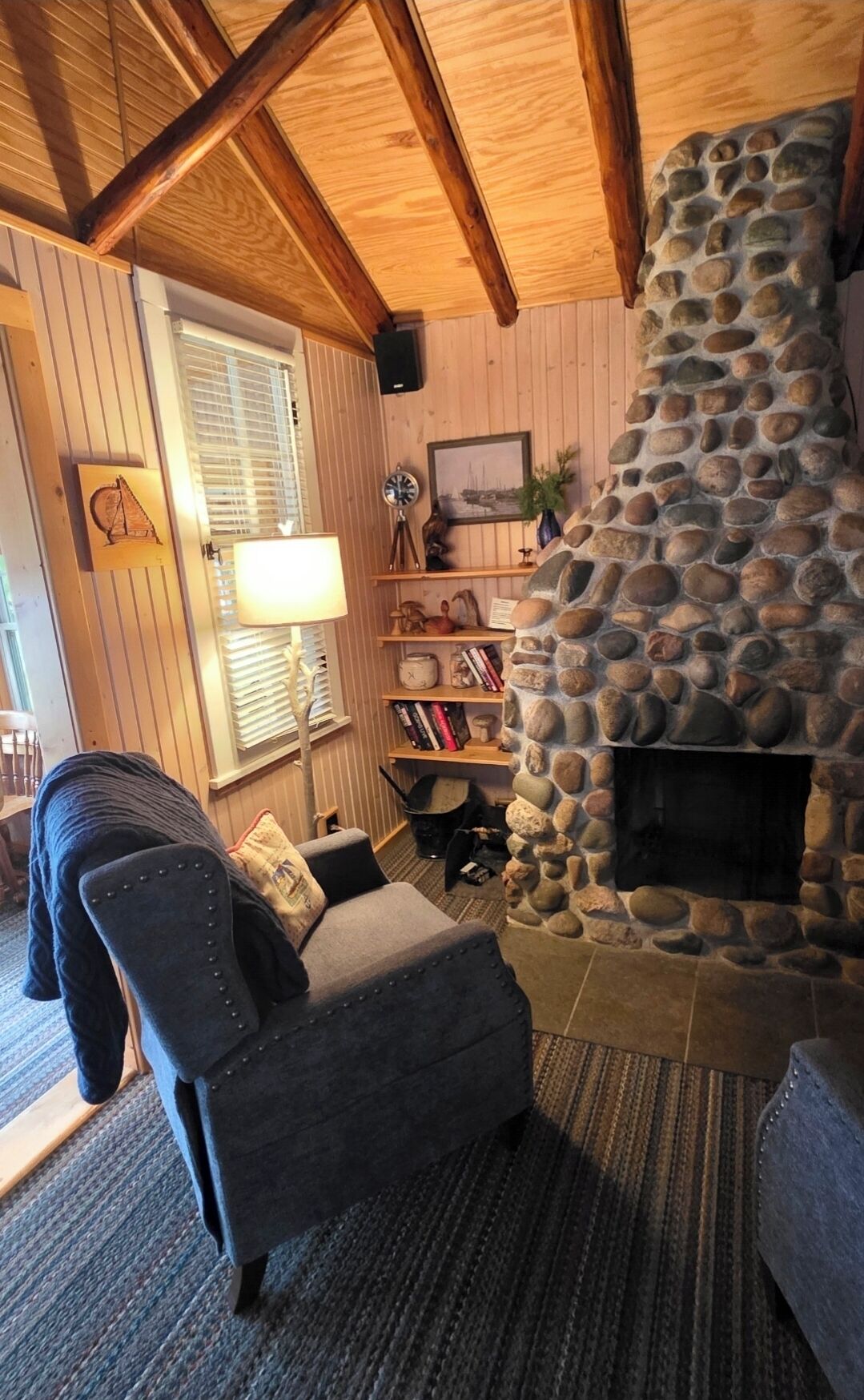A cozy living room with a stone fireplace, wooden beams, a comfortable armchair, and a bookshelf with various items.