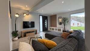 A modern living room with a large grey sofa, a wooden bench, a glass coffee table, and a flat-screen TV mounted on the wall. - IBARNIA - SUHESCUN - Gîte (SUHESCUN)