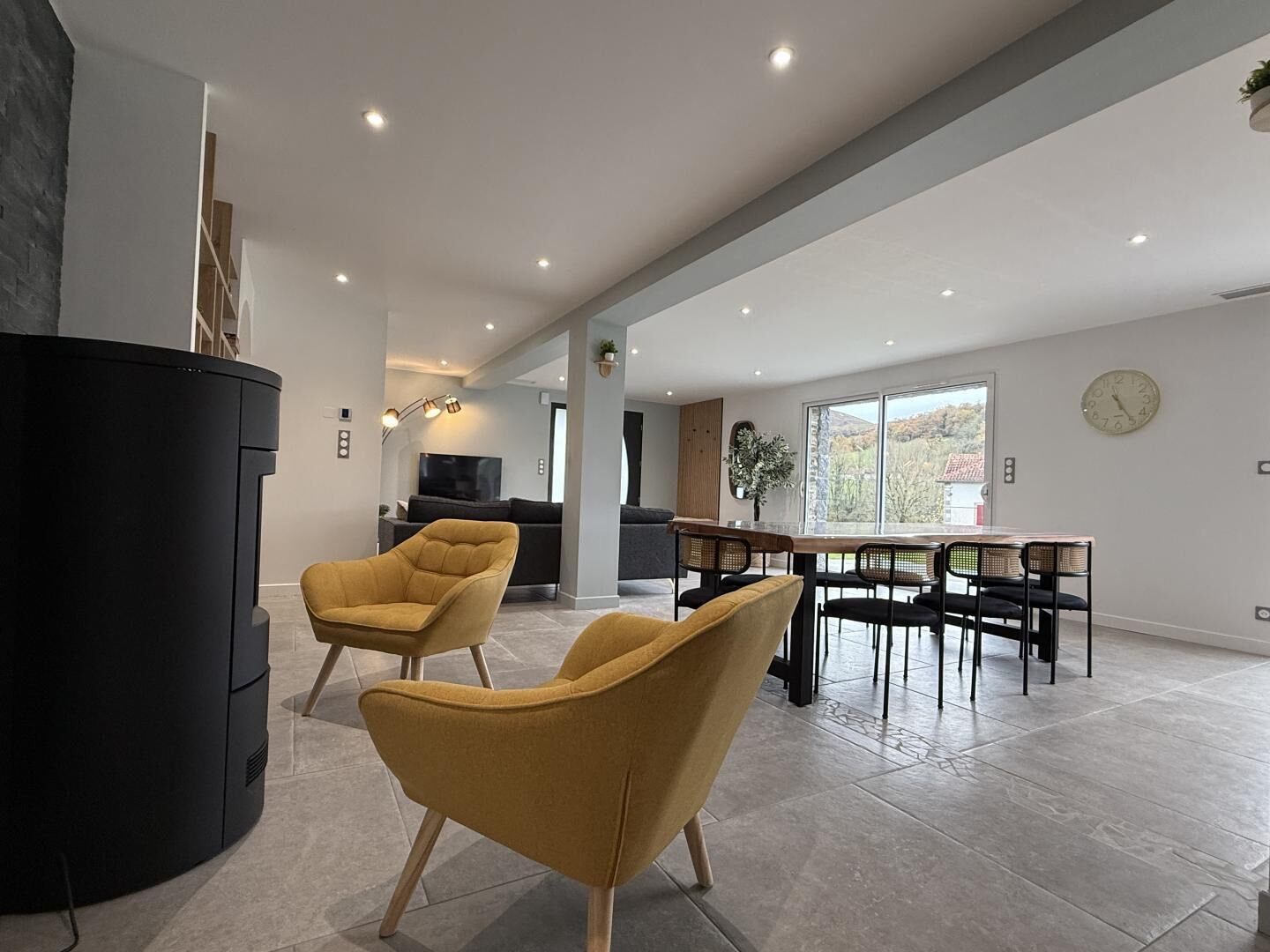 Een moderne woonkamer met een zwarte entertainmentkast, twee mosterdgele fauteuils, een eettafel met zwarte stoelen en een groot raam met uitzicht op de omgeving.