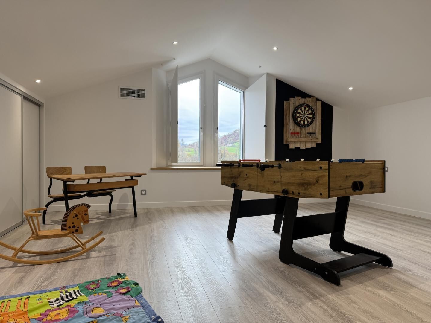 Een ruime kamer met een tafelvoetbalspel, een dartbord, een houten schommelpaard en een kleurrijk vloerkleed.