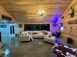 Un salon chaleureux avec un canapé d’angle, une table basse et un lustre. Un panneau est accroché au mur, sur lequel on peut lire : « Bienvenue à Neck of the Woods. »