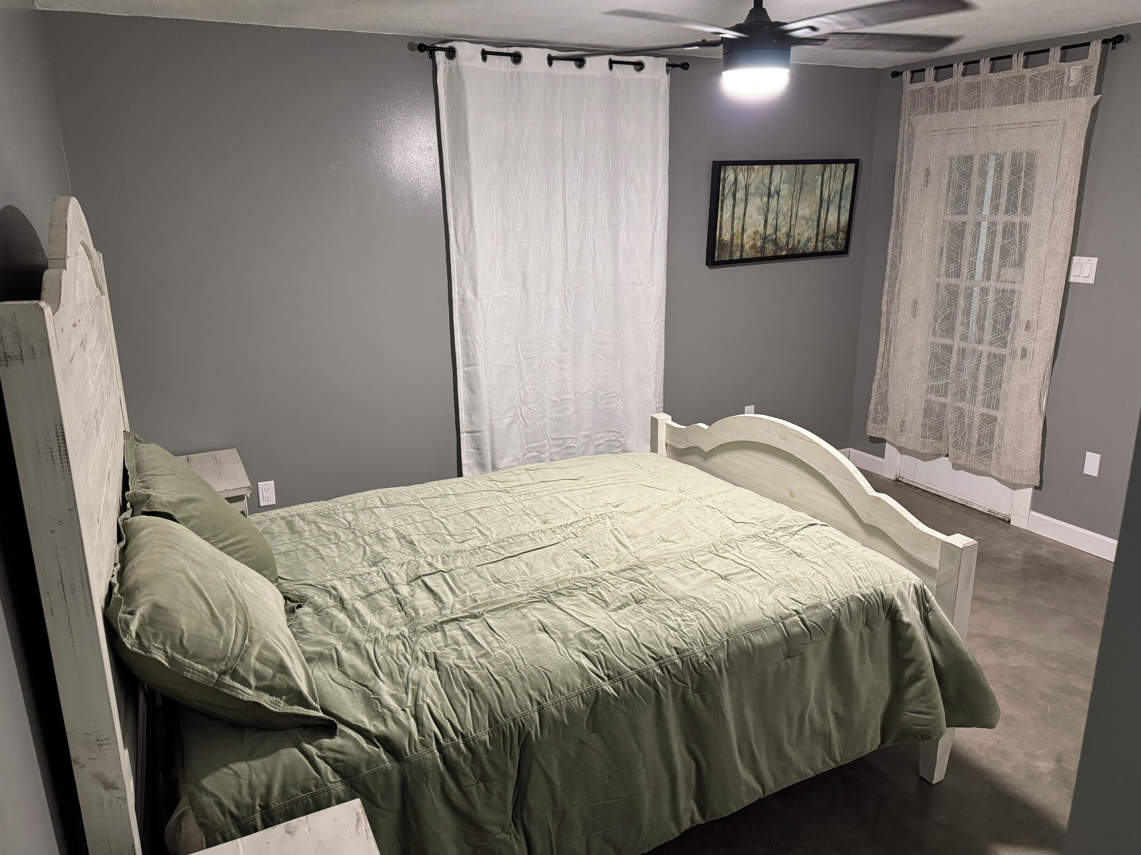 A bedroom with a bed, curtains, a ceiling fan, and a painting on the wall.