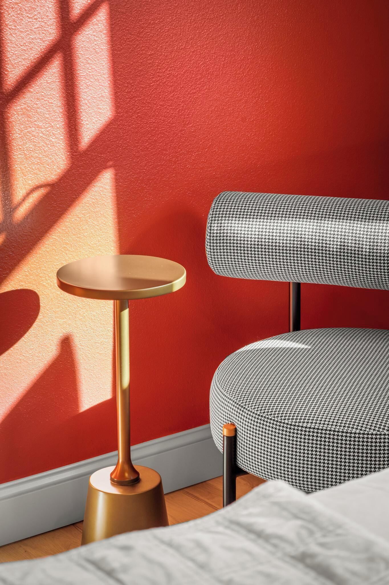 A modern room with a red wall, a round table with a gold base, and a houndstooth upholstered chair.