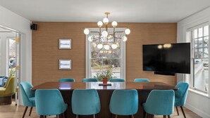 A modern dining area with a large wooden table, teal chairs, a chandelier, and a mounted TV. - Assemble Boise (Boise)