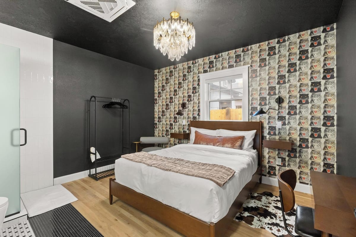 Un dormitorio con cama, candelabro, papel tapiz estampado, ventana y una pequeña mesa.