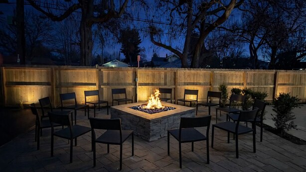 An outdoor patio with a stone fire pit surrounded by chairs, a wooden fence, and trees in the background. - Assemble Boise (Boise)