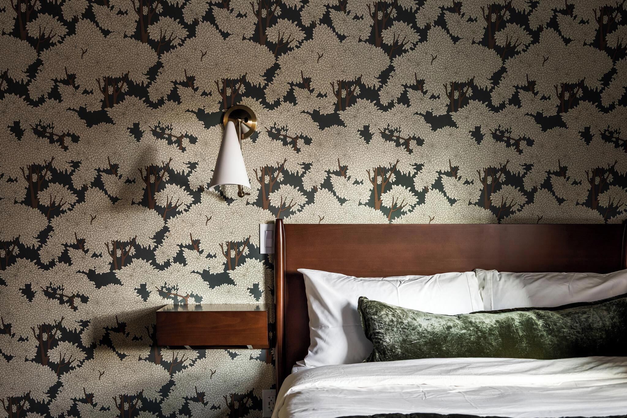 A bedroom with a bed, a nightstand, a lamp, and a wall with a tree pattern.