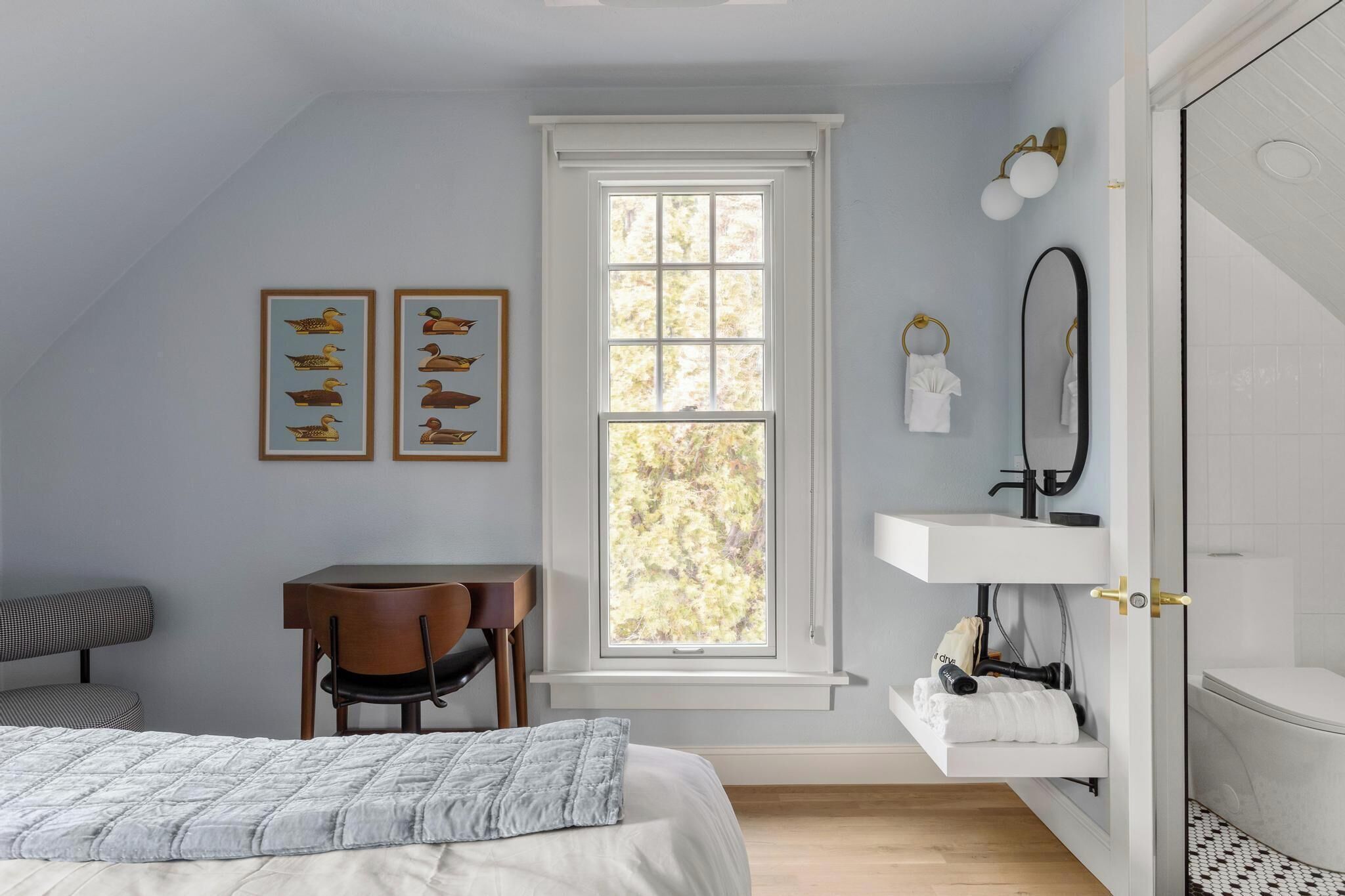 A bedroom with a bed, a desk, a chair, a window, a vanity with a mirror, and artwork on the wall.