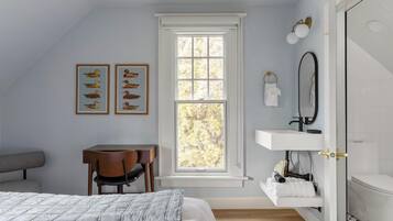 A bedroom with a bed, a desk, a chair, a window, a vanity with a mirror, and artwork on the wall.