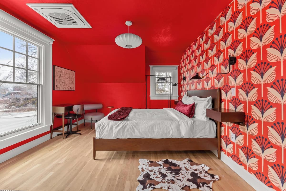 Un dormitorio con pared roja, cama con ropa de cama blanca, cabecero de madera y papel tapiz estampado.