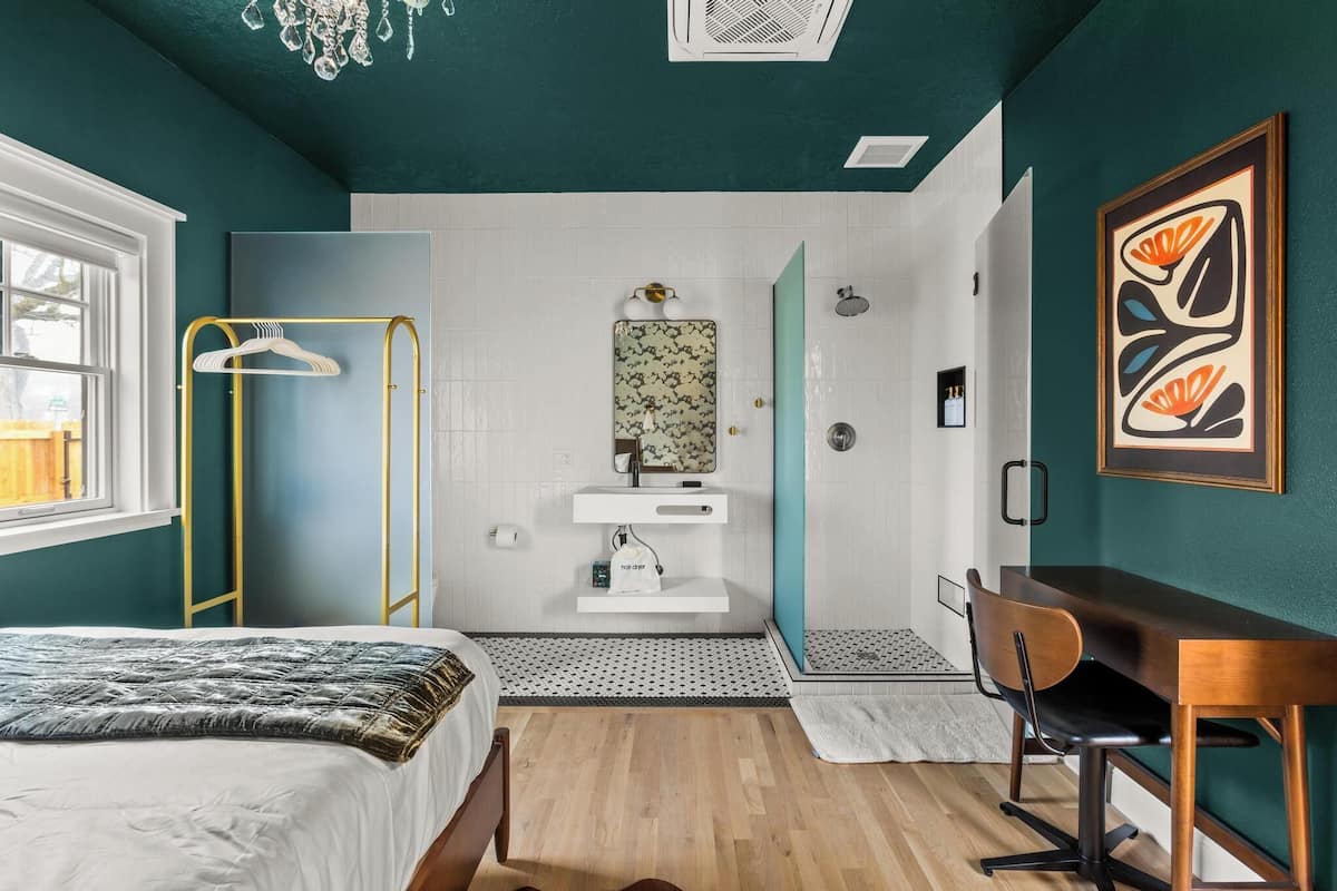 Un dormitorio con cama, escritorio, silla, armario, baño con lavamanos y espejo, y ducha.