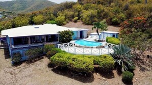 Une maison avec une piscine, entourée d’une clôture blanche, de verdure luxuriante et de collines.